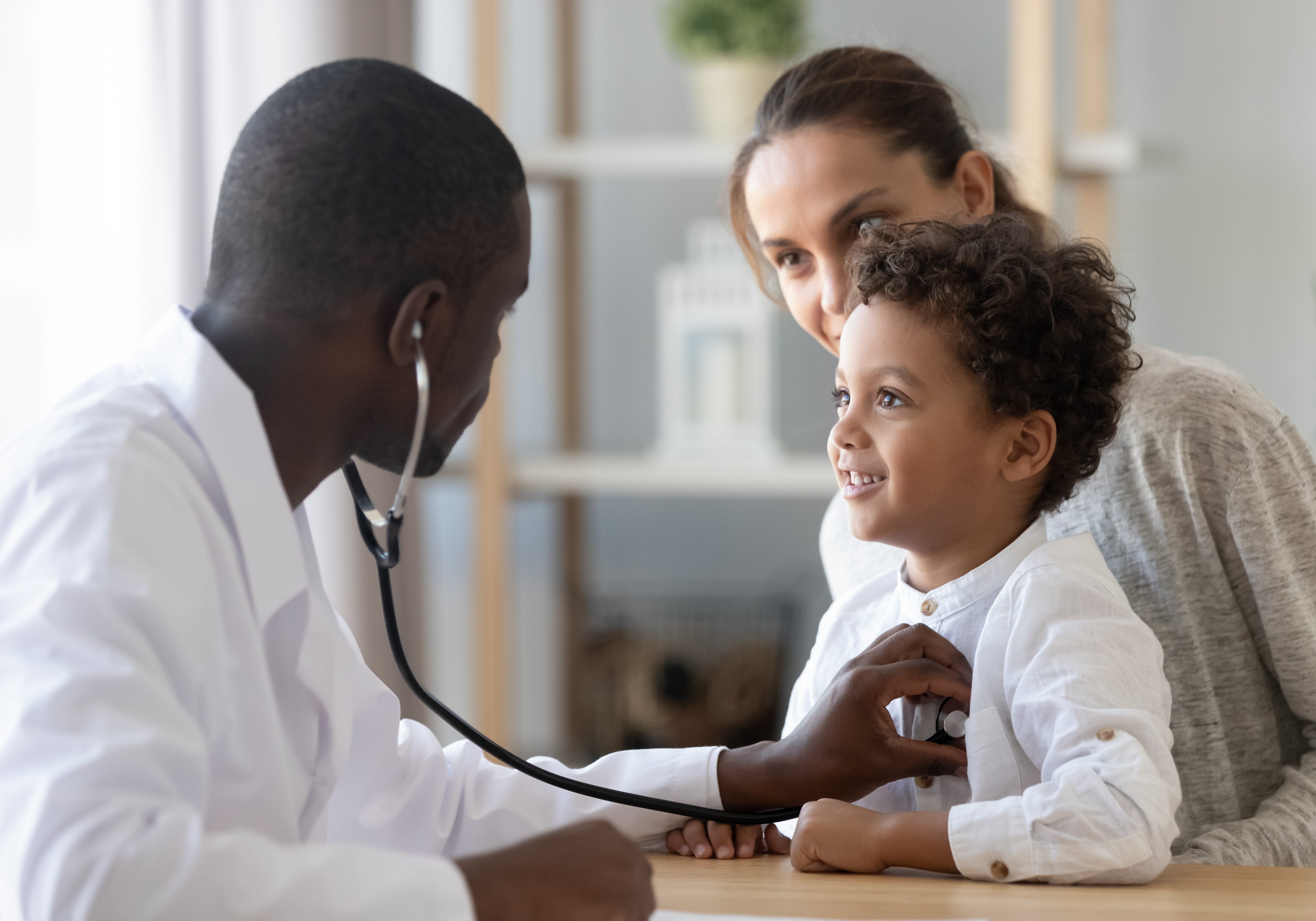 African American male pediatrician with stethoscope listening to lung and heart sound of little boy sitting on mother lap, physician checkup at home or in hospital, children medical insurance care